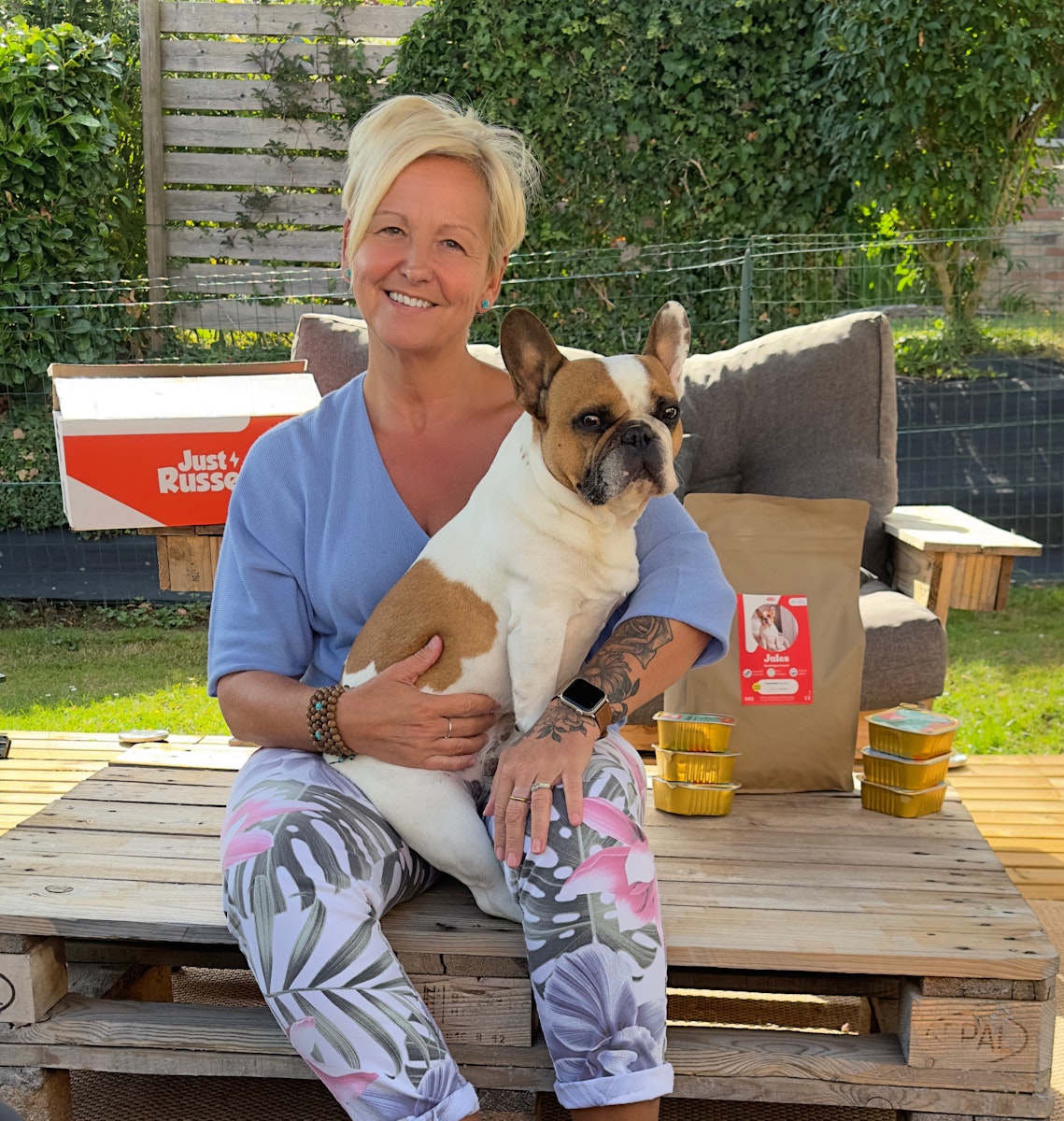 Woman holding a brown and white french bulldog outdoors on a wooden pallet with boxes and containers in the background.
