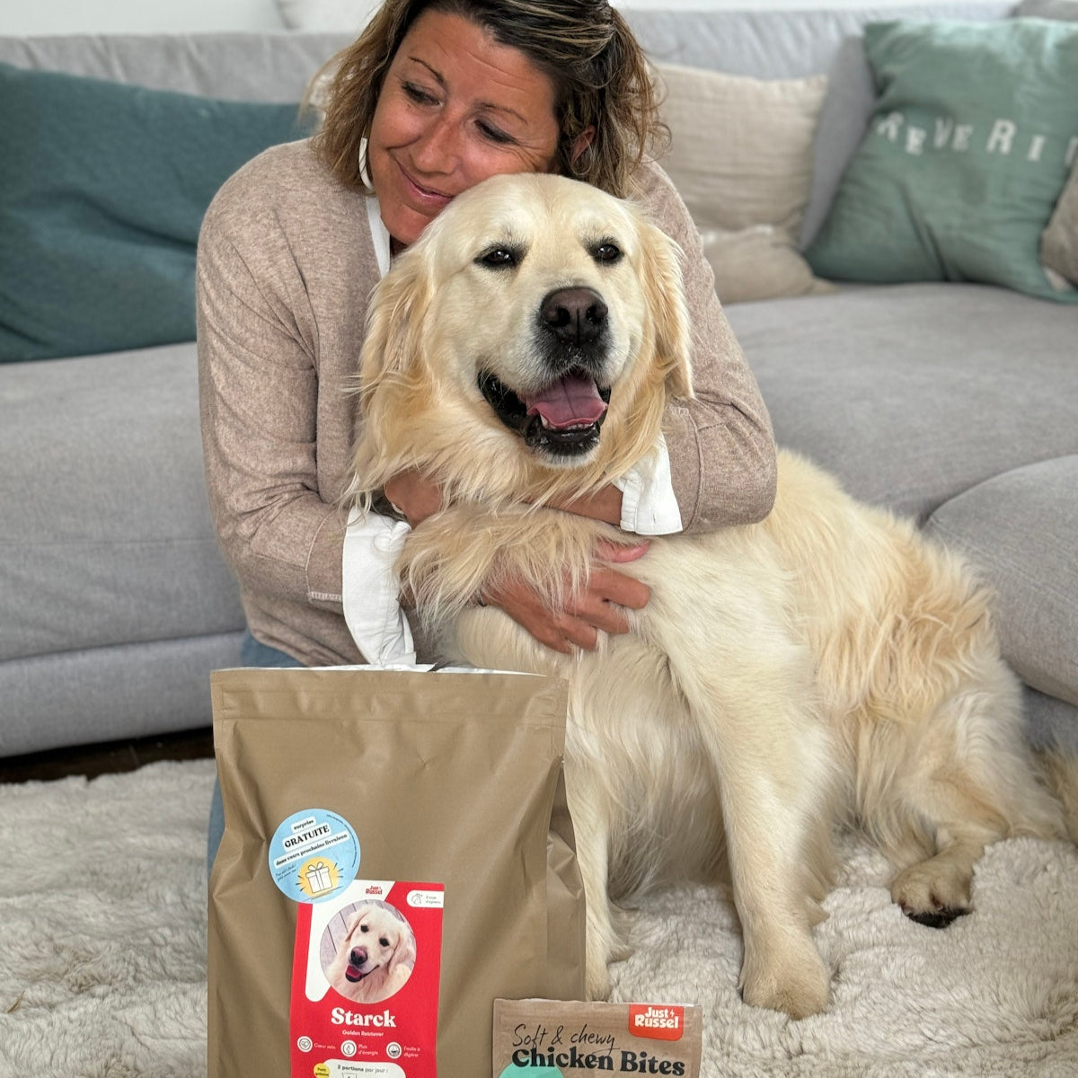 Woman with a golden retriever dog holding a bag of dog food on a carpeted floor.